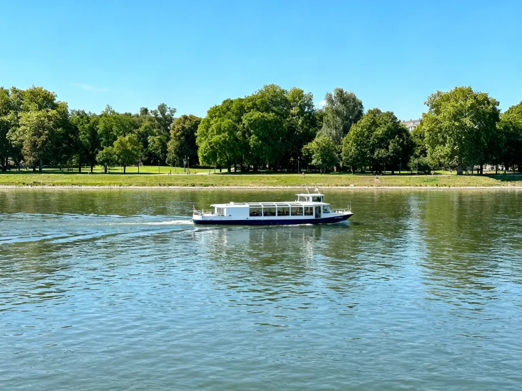 Mały prom pasażerski płynący przez rzekę Wisłę na tle zielonego brzegu parku w słoneczny dzień.